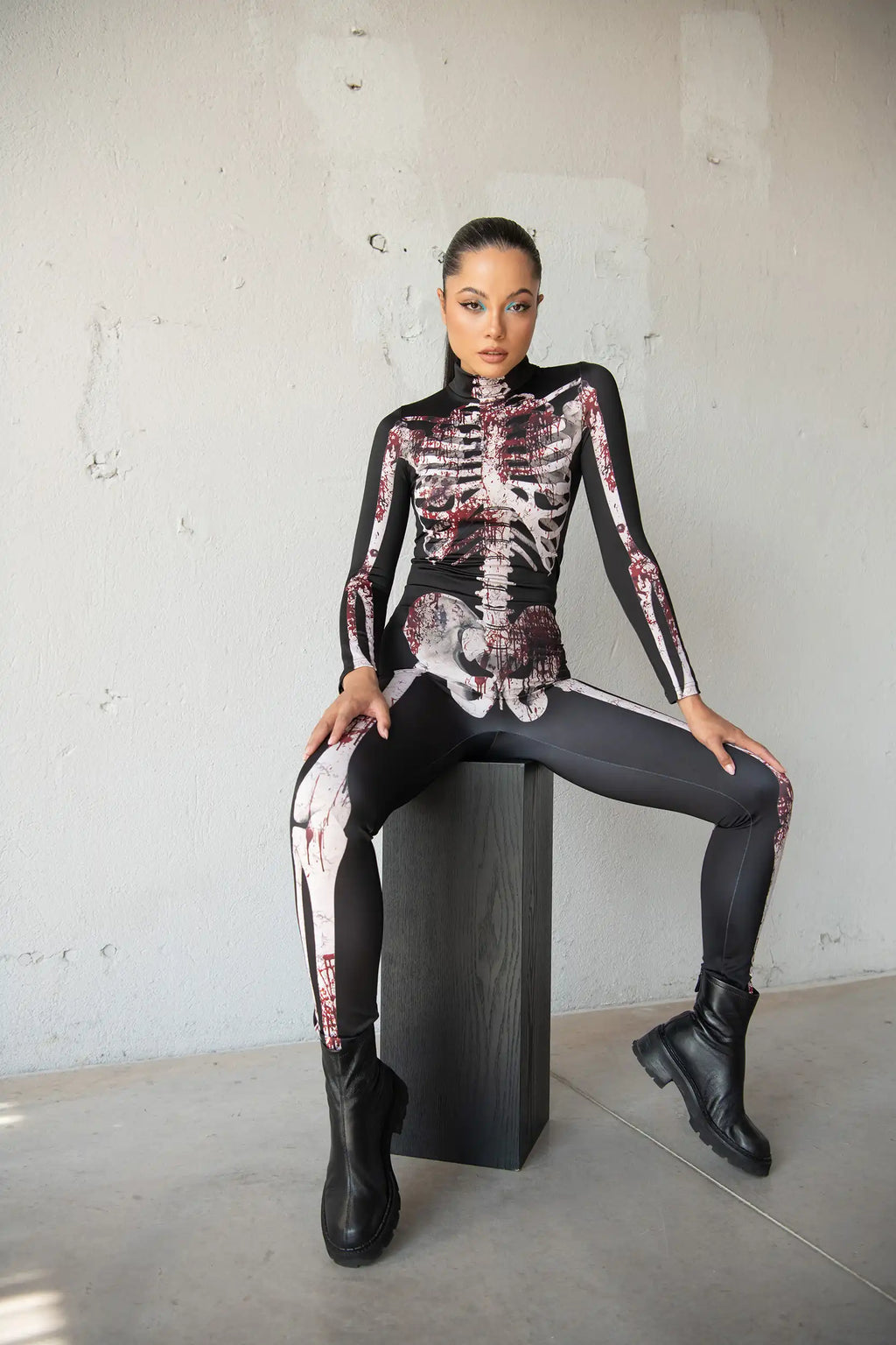 Person wearing a black skeleton-themed outfit sitting on a black box against a gray wall.