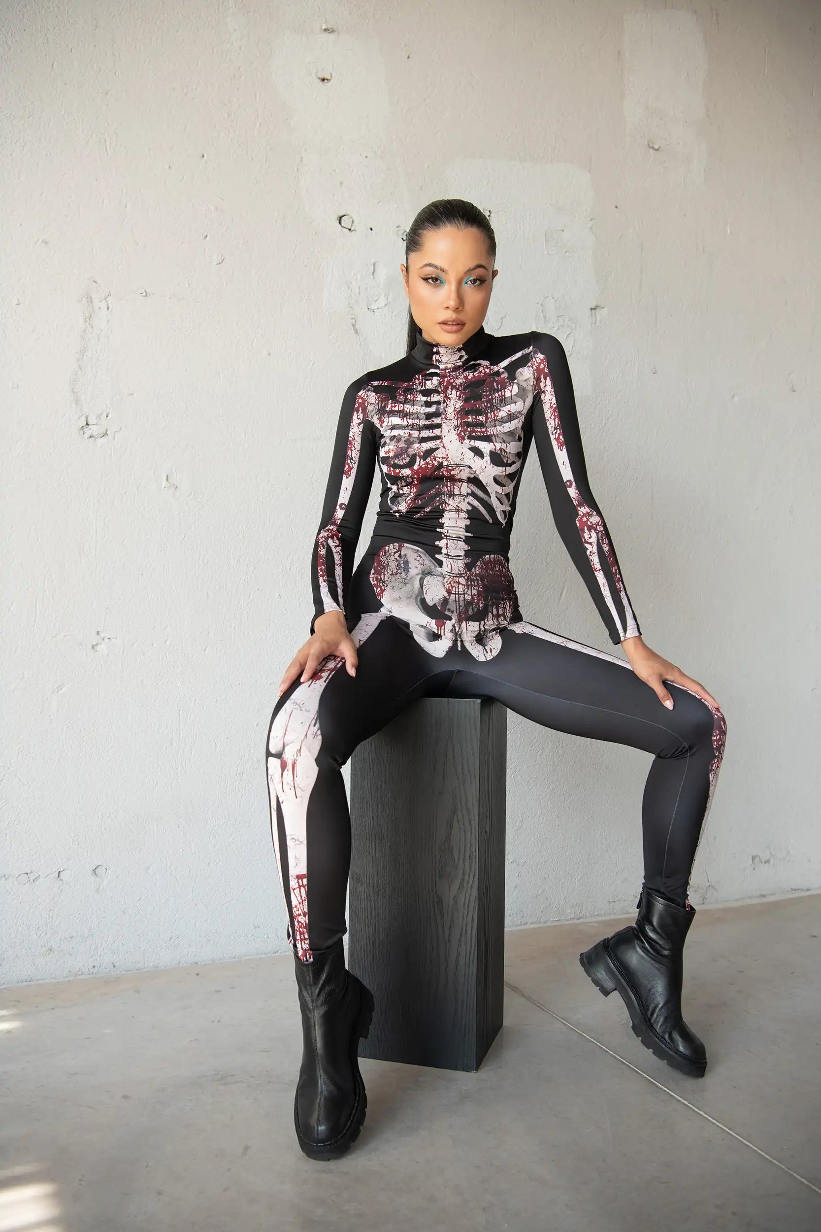 Person wearing a black skeleton-themed outfit sitting on a black box against a gray wall.